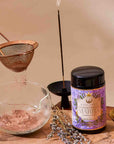 A jar of Flower Powder: Blueberry Lavender Matcha sits on a wooden board with a whisk, dried flowers, a teapot, cup of tea, sieve in use, and calming incense on a sunlit wooden table.