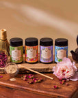 Four colorful tins of Flower Powder: Dragon Fruit Matcha are arranged on a wooden board with a bamboo whisk, scoop, teapot, rosebuds, matcha powder, and a pink peony for an elegant tea setup that celebrates tradition and gut health.