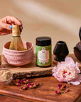 A hand whisks Flower Powder: Strawberry Matcha in a pink bowl on a wooden board, surrounded by matcha powder, a black teapot, flowers, rose petals, and glass jar—creating an inviting scene for mindful wellness support.