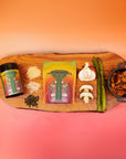 A wooden board displays Teasoning: Roasted Garlic Tea Salt, a spice jar, grated cheese, rice, peppercorns, mushrooms, asparagus, garlic, and a small pot of cooked potatoes—all set against an orange gradient background.