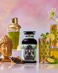 A decorative drink setup featuring the Summer Fridays: Wellness Mocktail Tea Bundle, with a gold Buddha head, cocktail shaker, ornate glasses of botanical tea mocktails, tea leaves, lime slices, and white flowers on a pink background.