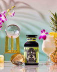 A tropical drink garnished with pineapple and an orchid sits beside a pineapple, a bottle labeled “Summer Fridays™: Pina Colada,” a tea infuser, orchids, and white flowers on a glossy surface with a pastel backdrop.