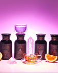 The Elemental Alchemy Master Collection features four dark glass bottles labeled Earth, Air, Water, Fire, a glowing pink crystal, a glass teacup with purple Elemental Alchemy tea, lemon and orange slices, set against a pink gradient background.