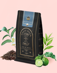 A bag of Earl Greater Grey: Tea for the Bright & Bold rests on a pink background, surrounded by tea leaves, bergamot, and green foliage. The packaging showcases gold celestial-themed graphics and elegant labeling.