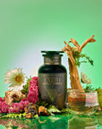 A dark green bottle labeled "Earth: Stabilizing and Nourishing Herbs, Roots, and Puerh tea" sits with flowers, bark, moss, a glass teacup of grounding tea, and a branch on a glossy surface against a green background for immune support.