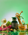 A dark green "Earth: Stabilizing and Nourishing Herbs, Roots, and Puerh tea" bottle sits among flowers, moss, wood, and herbs. Next to it is a clear cup of golden tea against a fresh green gradient background.