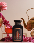 An arrangement of elegant tea-making items includes a tin labeled "The Magician" tea, a glass teacup filled with tea, various pink and red flowers, a gold teapot, and a crystal ball. The scene is set against a neutral backdrop, creating a sophisticated aesthetic.