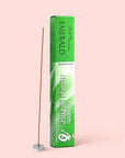 A green box labeled “Natural Incense : Emerald” stands upright next to a lit sandalwood incense stick in a small white holder, all set against a pale pink background.