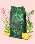 A green geometric tea bag package of Emerald - Cherry Blossom Pineapple Cream Green Tea is surrounded by pink cherry blossoms, tea leaves, and pineapple slices, set against a pale pink background.