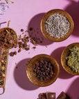An overhead view of four ornate brass bowls on a pink surface. Each bowl contains different organic herbs and spices. The bowls are arranged neatly, casting shadows around them. Vibrant flowers and chocolate pieces surround the bowls, adding colorful touches reminiscent of a Magic Hour's The Empress: Lavender Currant Shatavari Cocoa Tea of Nurturing Creativity setup.