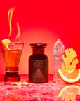 A vibrant red scene displays Fire—a cocktail with a lemon twist emitting flames, a dark "Fire" bottle, metabolism-supporting herbs, cardamom pods, ginger root, quartz crystal, Fire Tea, and orange on marble.