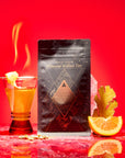 A glass of tea with lemon sits next to a black Magic Hour Fire package. Ginger, an orange slice, and a pink crystal rest on red marble, highlighting this metabolism-boosting herbal blend against a bold red background.