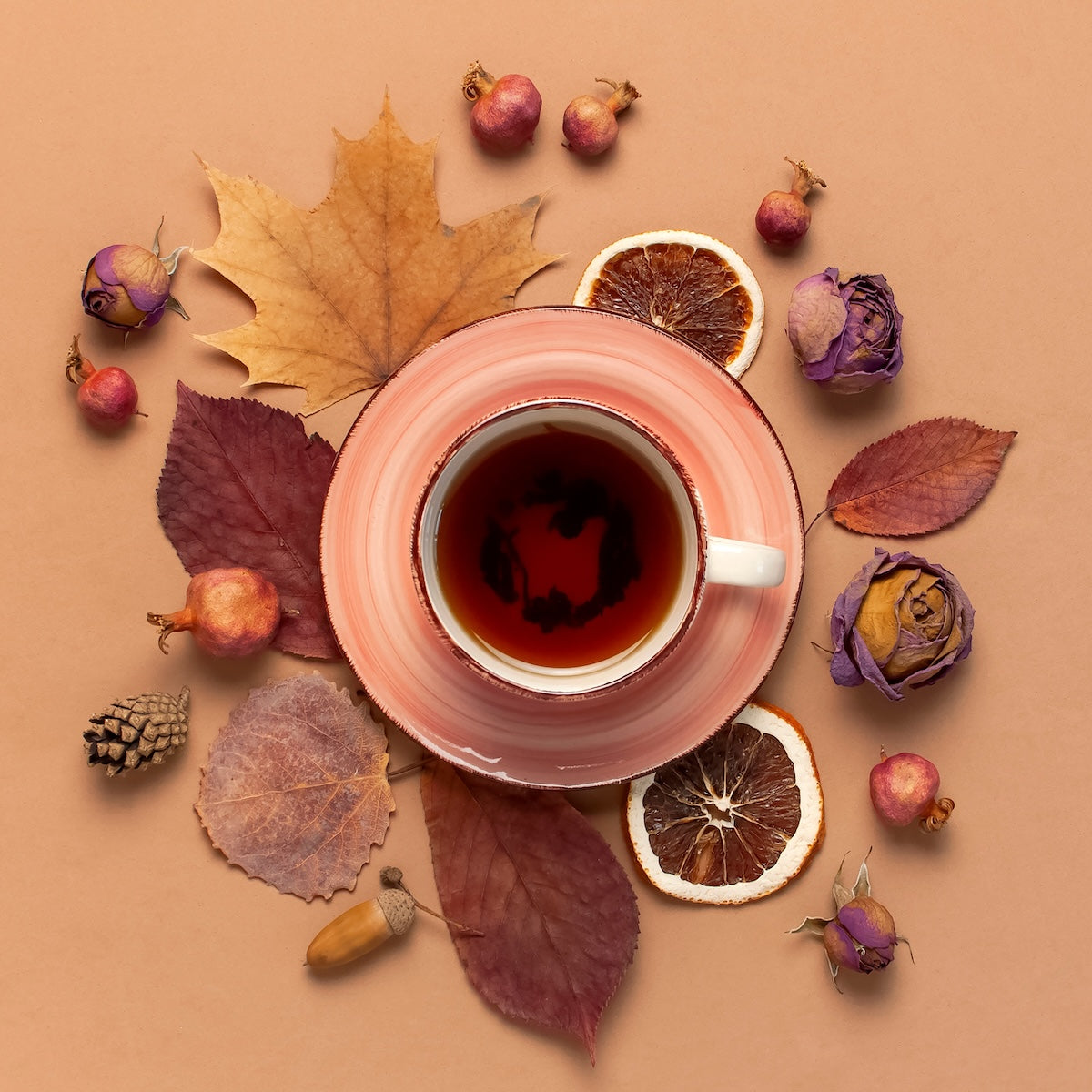 A cup of tea in a pink saucer is surrounded by dried leaves, orange slices, small pinecones, acorns, and dried flowers on a beige background, creating an autumnal arrangement reminiscent of the Fall Flavors Tea Sampler Set.