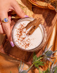 A hand holds a glass of creamy spiced chai, garnished with cinnamon sticks, resting on a wooden board. The scene is enriched by the Fall Flavors Tea Sampler Set, featuring an array of dried leaves and purple thistle flowers that enhance the warm autumn ambiance against the golden surface.