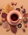 A cup of tea in a pink saucer is surrounded by dried leaves, orange slices, small pinecones, acorns, and dried flowers on a beige background, creating an autumnal arrangement reminiscent of the Fall Flavors Tea Sampler Set.