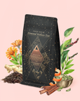 A black and gold Fire: Spiced Black Tea with Ginger and Cardamom to Ignite Courage and Vitality package is surrounded by botanical elements like ginger, cinnamon, orange peel, flowers, and green leaves on a light pink background. This adaptogenic tea blend features metabolism-boosting herbs designed to invigorate your senses and support courage and vitality.
