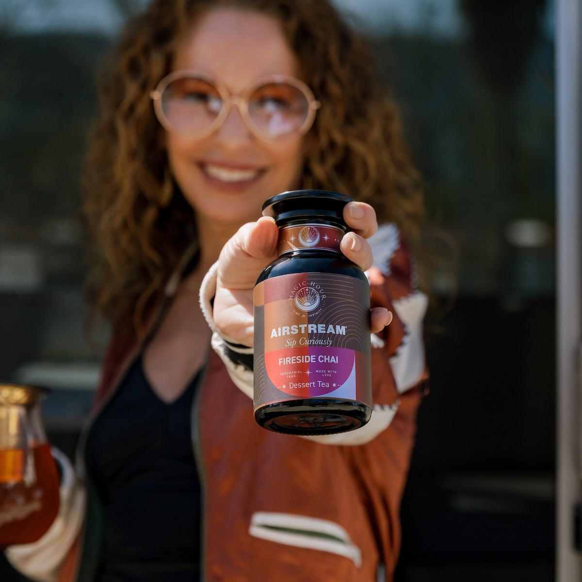 Zhena smiling in the background holding a violet glass apothecary jar of Fireside Chai herbal chai loose leaf tea.