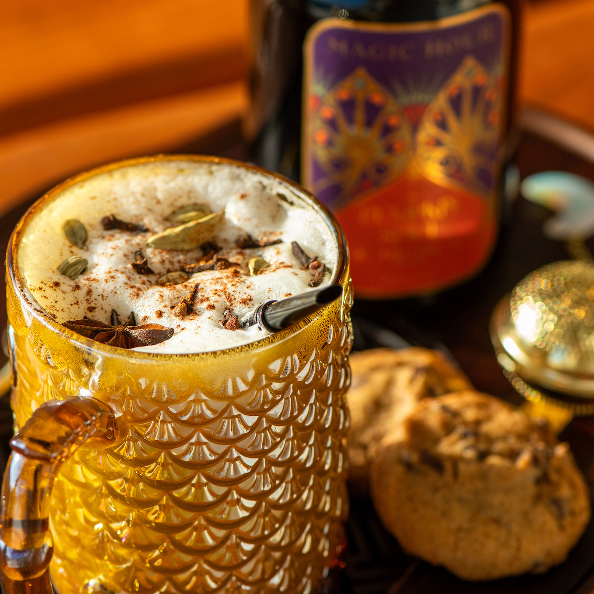 A warm beverage topped with frothy milk and spices is served in a textured golden glass mug. Beside it are two cookies on a wooden tray. A blurred bottle with vibrant labeling is in the background.
