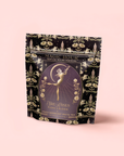 A decorative black and gold tea package labeled "Flora In Bloom: Peach Moonlight White Tea" by Magic Hour features an elegant, celestial feminine figure amid floral and star motifs on a light pink background.