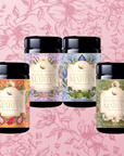 Four colorful Flower Powder: Matcha Bundle canisters with floral labels are displayed side by side on a pink background with illustrated flowers and leaves. Each features a unique matcha flavor, such as Strawberry Matcha and Blueberry Lavender Matcha.