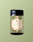 A black jar of Flower Powder: Strawberry Matcha, made with organic ceremonial matcha, features a floral and strawberry-themed label against a solid olive green background.