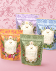 Four colorful pouches from the Flower Powder: Matcha Bundle are arranged in front of a pink floral wallpaper, each labeled with a different flavor—Dragon Fruit, Strawberry, Berry Lavender, and Berry Bergamot. Crafted with antioxidant-rich Kagoshima ceremonial matcha, these vibrant blends deliver both exquisite taste and wellness benefits in every cup.