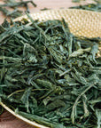 Loose dried Flower Fragrance Iccha Kariban Japanese green tea leaves on a textured golden plate, with some leaves scattered on a wooden surface in the background.