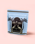 A light blue Magic Hour "French 75" Lemon Sparkling Wine White Mocktail Tea package with peacocks, palms, and gold-black details stands against a pale pink background—perfect for a wellness mocktail with Silver Needle white tea.
