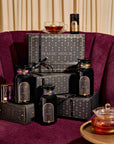 A velvet chair showcases a beautifully curated Branded Gift Box alongside Magic Hour tea boxes and black tea jars, with a teacup perched on top and a glass teapot labeled Magic Hour elegantly resting on a gold tray beside it. The scene is set against cream-colored curtains, creating an inviting environment perfect for an elevated gifting experience.