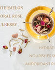 A cup of tea infused with various floral ingredients, including rose petals and dried mulberries, sits next to some scattered tea leaves and flowers. The words "WATERMELON, FLORAL ROSE, MULBERRY" and "HYDRATING, NOURISHES SKIN, ANTIOXIDANT RICH" are displayed around this organic loose leaf tea from Magic Hour called Gemini: Watermelon-Rose-Mulberry Pomegranate Tea for Beauty, Balance & Quenching Curiositeas.