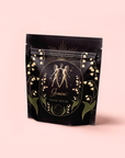 A black package with gold and green art, mirrored figures, "Gemini" text, florals, and a pink label that reads "Magic Hour," highlighting Gemini: Watermelon-Rose-Mulberry Pomegranate Tea for Beauty, Balance & Quenching Curiositeas.