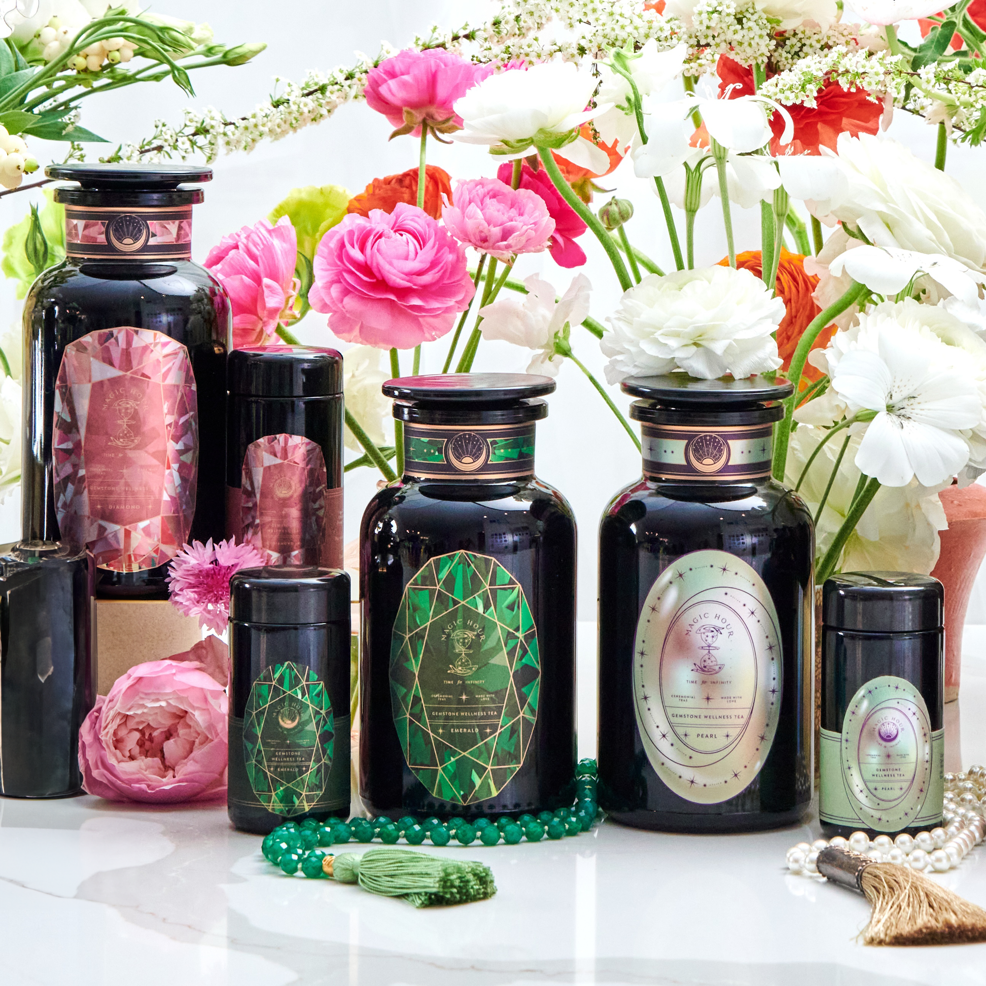 A display of dark glass jars and containers with ornate labels featuring green and pink designs, surrounded by pink and white flowers. Some containers are cylindrical, hinting at treasures of the Magic Hour Gemstone Bundle with Diamond, Emerald, and Pearl, with a green beaded necklace and a tassel in the foreground.