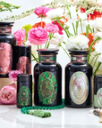 A display of dark glass jars and containers with ornate labels featuring green and pink designs, surrounded by pink and white flowers. Some containers are cylindrical, hinting at treasures of the Magic Hour Gemstone Bundle with Diamond, Emerald, and Pearl, with a green beaded necklace and a tassel in the foreground.