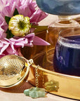 A gold Emerald Star Tea Strainer rests on a shiny gold tray alongside purple and white flowers and a glass of blue liquid. In the background, there is another blue drink in a glass, and two small stones are placed next to the tea strainer.