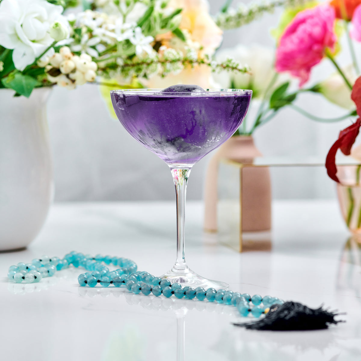 A vibrant purple cocktail in a coupe glass sits on a white surface, surrounded by a dazzling piece from Magic Hour's Gemstone Sampler Tea Set and floral arrangements in the background. The drink's vivid color contrasts with the delicate white and pink flowers, creating an elegant and colorful scene.