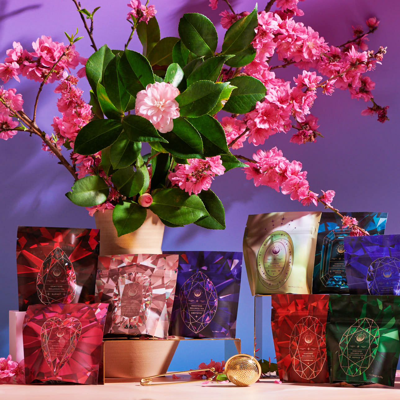 A display of colorful tea packages from the Magic Hour's Gemstone Sampler Tea Set is arranged in front of a vibrant purple backdrop, adorned with green leaves and pink blossoms. The packages feature various geometric designs, and there is a gold tea infuser placed among the bags of adaptogens and gemstone teas.