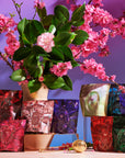 A display of colorful tea packages from the Magic Hour's Gemstone Sampler Tea Set is arranged in front of a vibrant purple backdrop, adorned with green leaves and pink blossoms. The packages feature various geometric designs, and there is a gold tea infuser placed among the bags of adaptogens and gemstone teas.