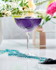 A vibrant purple cocktail in a coupe glass sits on a white surface, surrounded by a dazzling piece from Magic Hour's Gemstone Sampler Tea Set and floral arrangements in the background. The drink's vivid color contrasts with the delicate white and pink flowers, creating an elegant and colorful scene.