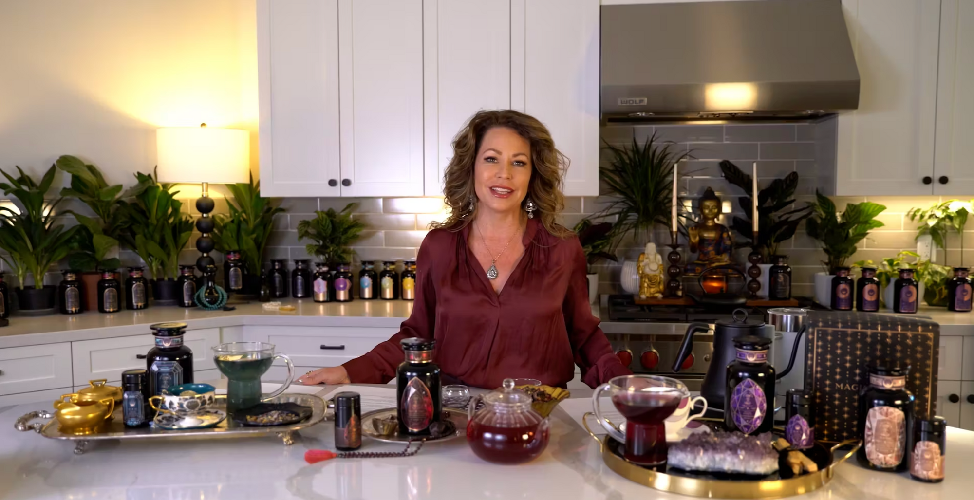 A woman stands in a modern kitchen with grey backsplash and white cabinetry, surrounded by various ornate bottles, gemstones, and candles. She wears a maroon blouse and jewelry. Green plants and a Buddha statue in the background enhance the serene ambiance.