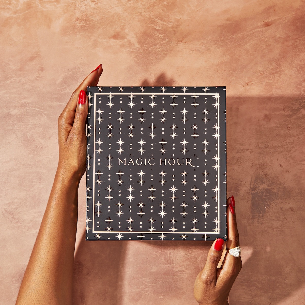 A person with red nail polish and a ring holds a dark blue, star-patterned box labeled "Magic Hour: The Goddess of Earl Set" against a peach-colored textured background.