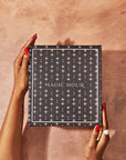 A person with red nail polish and a ring holds a dark blue, star-patterned box labeled "Magic Hour: The Goddess of Earl Set" against a peach-colored textured background.