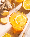 A glass of Ginger Lemon with lemon slices, surrounded by fresh ginger root, ginger slices, and lemon halves on a light surface. A glass straw is in the drink.