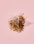 A pyramid-shaped green tea sachet of Tea for Abundance: Goddess Green: Apricot & Lychee Green Tea with organic lemongrass resting on a pile of loose-leaf tea with a small circular tag.