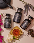 The Goddess of Earl: Lavender London Fog—Tea for Blooming Clarity & Calm Moods sits in a labeled jar beside a glass teacup, loose tea leaves, a gold and black teapot, and flowers including an orchid and rose on a light surface.