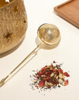 A Gold Luxe Loose Leaf Tea Strainer with a long handle sits alongside a small pile of colorful petal and leaf-infused loose-leaf tea. These items are arranged on a white surface beside The Goddess of Earl Set by Magic Hour, which includes a textured, gold-colored container featuring star-shaped cutouts and a wooden tray.