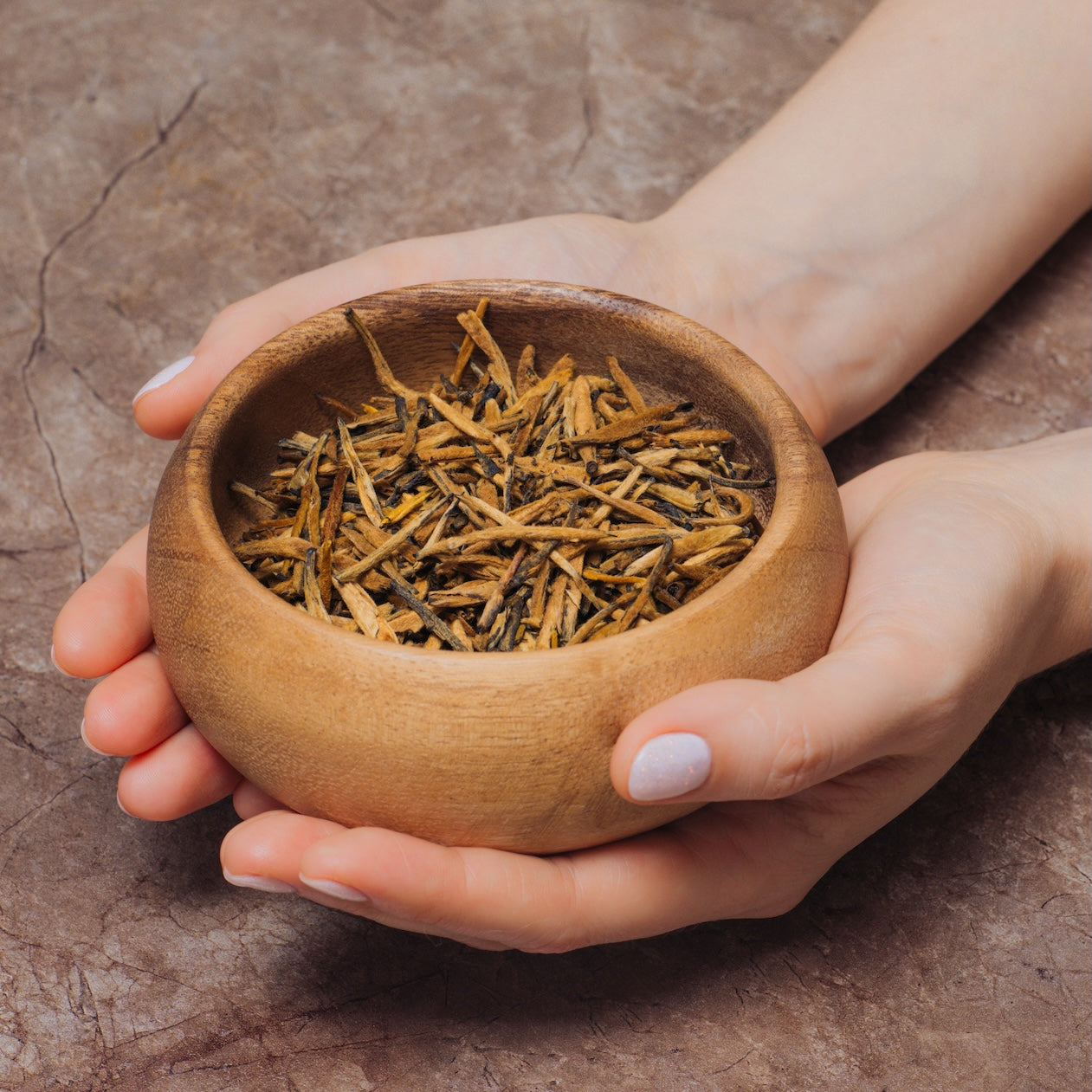 A pair of hands gently holds a round wooden bowl filled with Yunnan Gold Buds Black Tea on a textured brown surface.