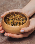 A pair of hands gently holds a round wooden bowl filled with Yunnan Gold Buds Black Tea on a textured brown surface.