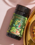 A black cylindrical container labeled "Green Grape Yerba Mate: Cosmic Garden Iced Tea" from Magic Hour is placed next to a gold dish filled with organic loose leaf tea leaves and herbs. The backdrop is a light pink surface with shadows adding depth.