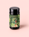 A black tea canister with a colorful floral label reading “Green Grape Yerba Mate: Cosmic Garden Iced Tea” sits on a light pink background. The decorative label is adorned with fruits, flowers, and leaves, suggesting vibrant ingredients like spirulina and green yerba mate woven into the design.