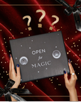 A person with dark blue nails holds the Black Friday Mystery Box labeled "OPEN for MAGIC" above red velvet, gold confetti, and black ribbons, with three gold question marks overhead—suggesting premium teas or tea accessories inside.